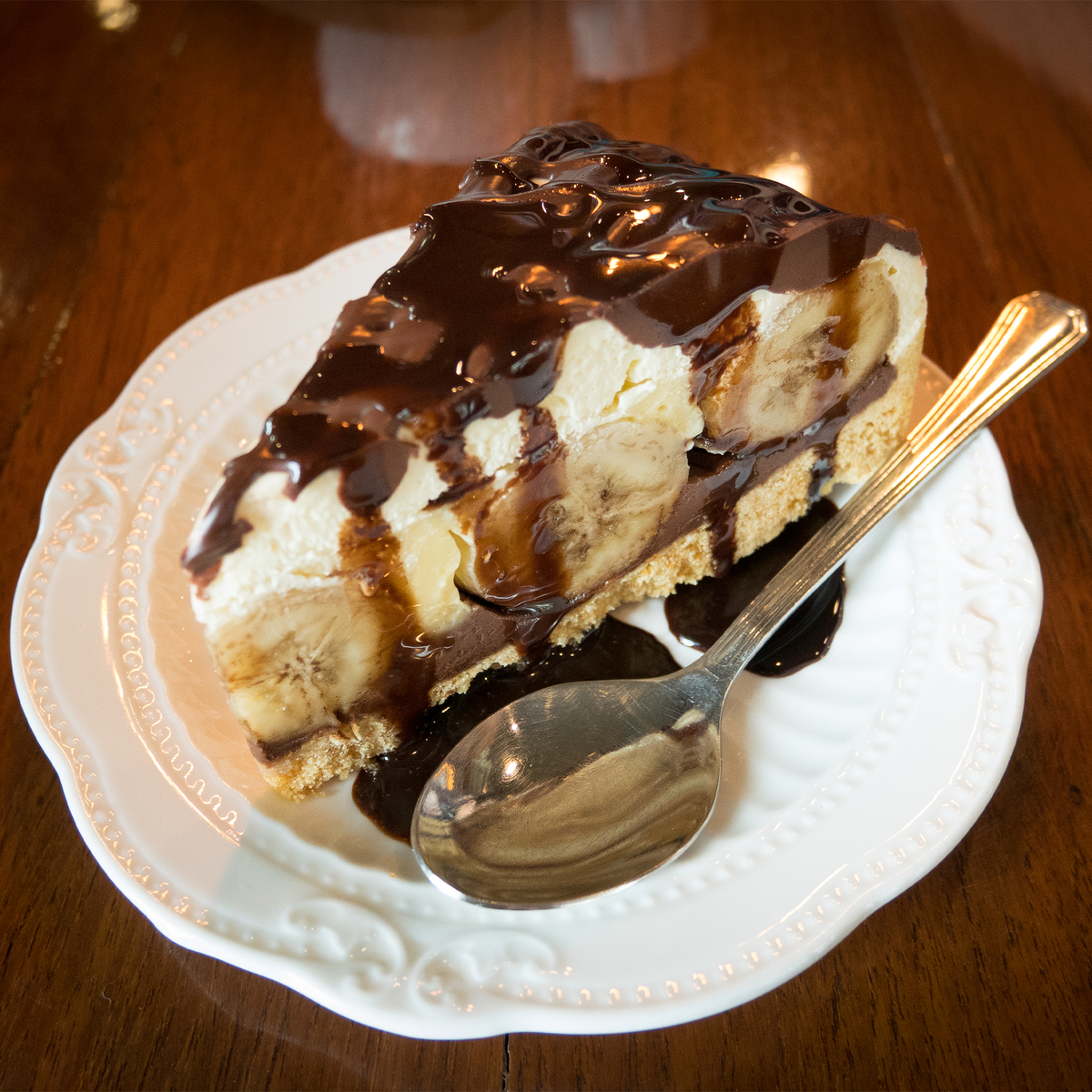 A slice of vanilla banana pie dessert topped with chocolate sauce and caramel drizzle, served on a white plate with a spoon on a wooden table.