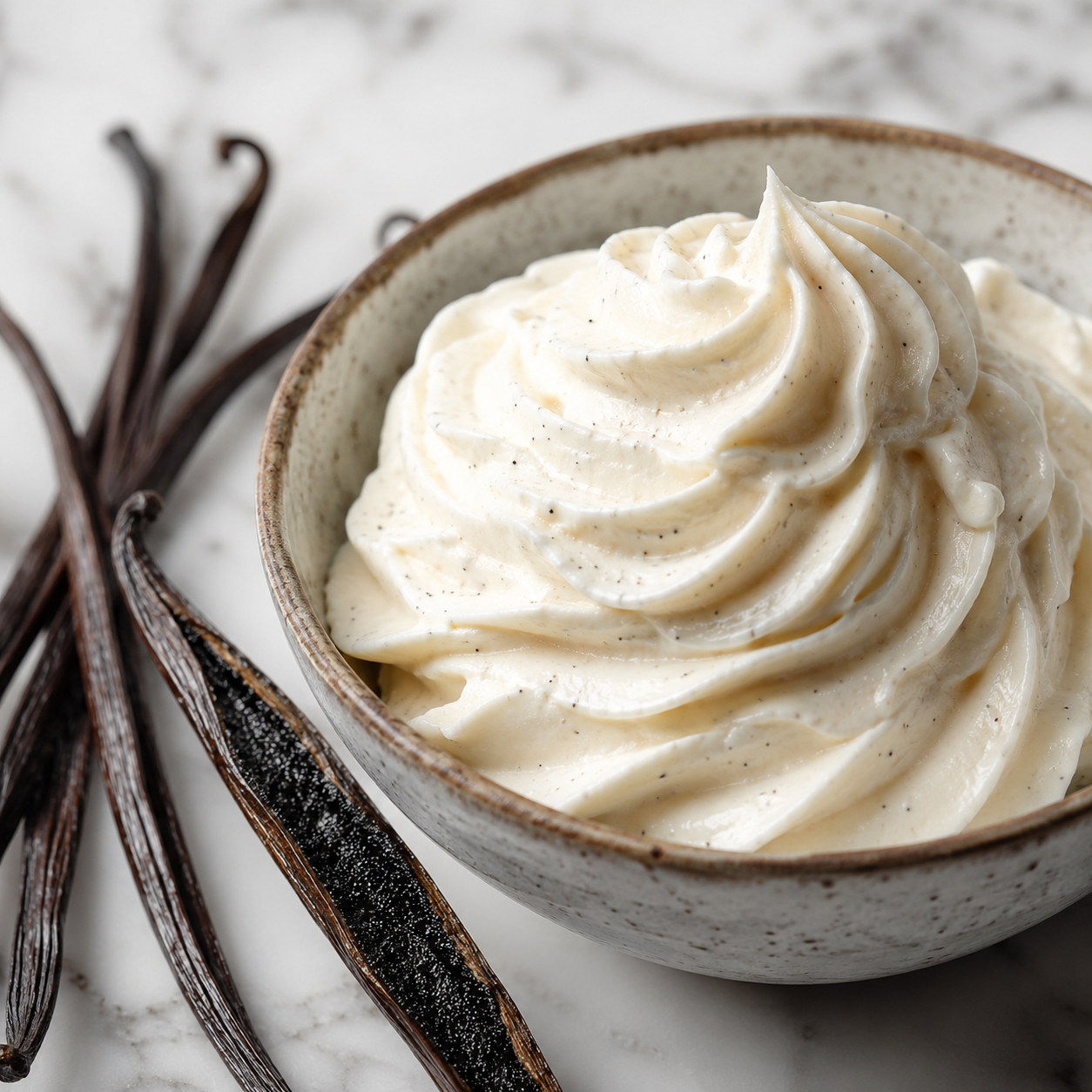 A close-up of whipped cream or vanilla ice cream swirled in a speckled ceramic bowl, with vanilla beans placed beside it on a light marble surface.