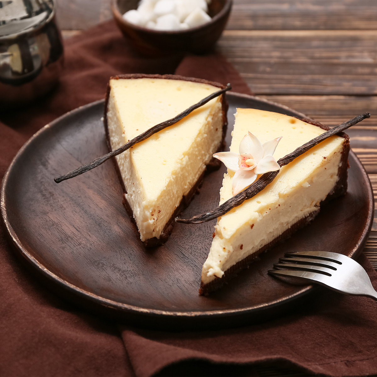 Two slices of creamy cheesecake on a dark brown ceramic plate, garnished with vanilla beans and a small flower, set on a rustic wooden table with a fork nearby.