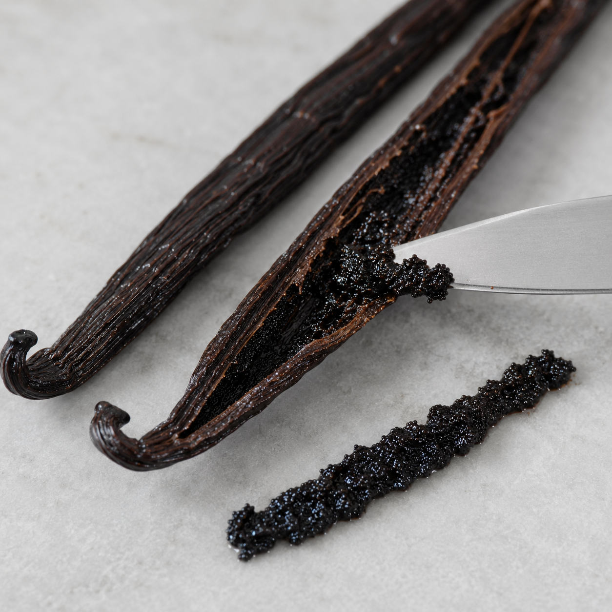 Two dark vanilla beans on a light stone surface, one opened with a silver knife and a small smear of dark vanilla paste beside it.