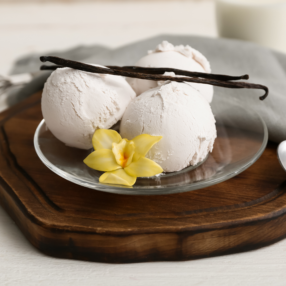 Three scoops of vanilla ice cream on a clear glass plate, garnished with two vanilla beans and a small yellow flower, set on a wooden board.