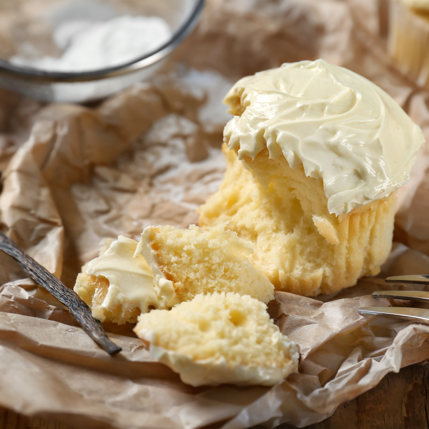 A close-up of a vanilla sponge cake muffin with thick white frosting, shown with a slice cut out on crumpled brown paper. A vanilla bean and a fork are visible in the scene.