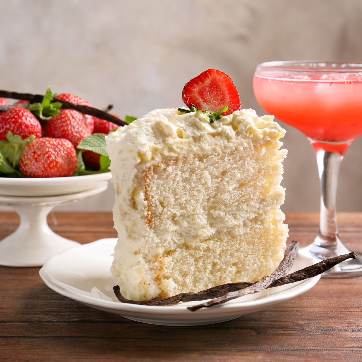 A slice of white cake on a white plate, garnished with a strawberry slice, set on a wooden table with vanilla beans and a pink cocktail in the background.