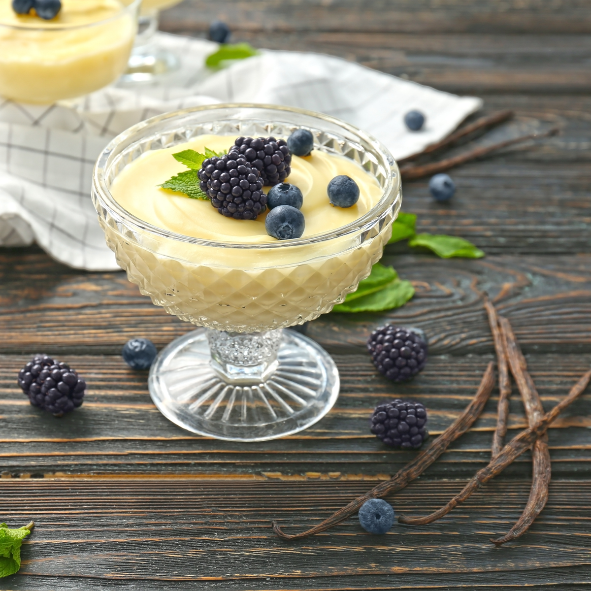A glass dessert cup filled with pale yellow vanilla custard, topped with fresh blueberries, blackberries, and mint leaves, set on a rustic dark wood table with vanilla beans and additional berries scattered around.