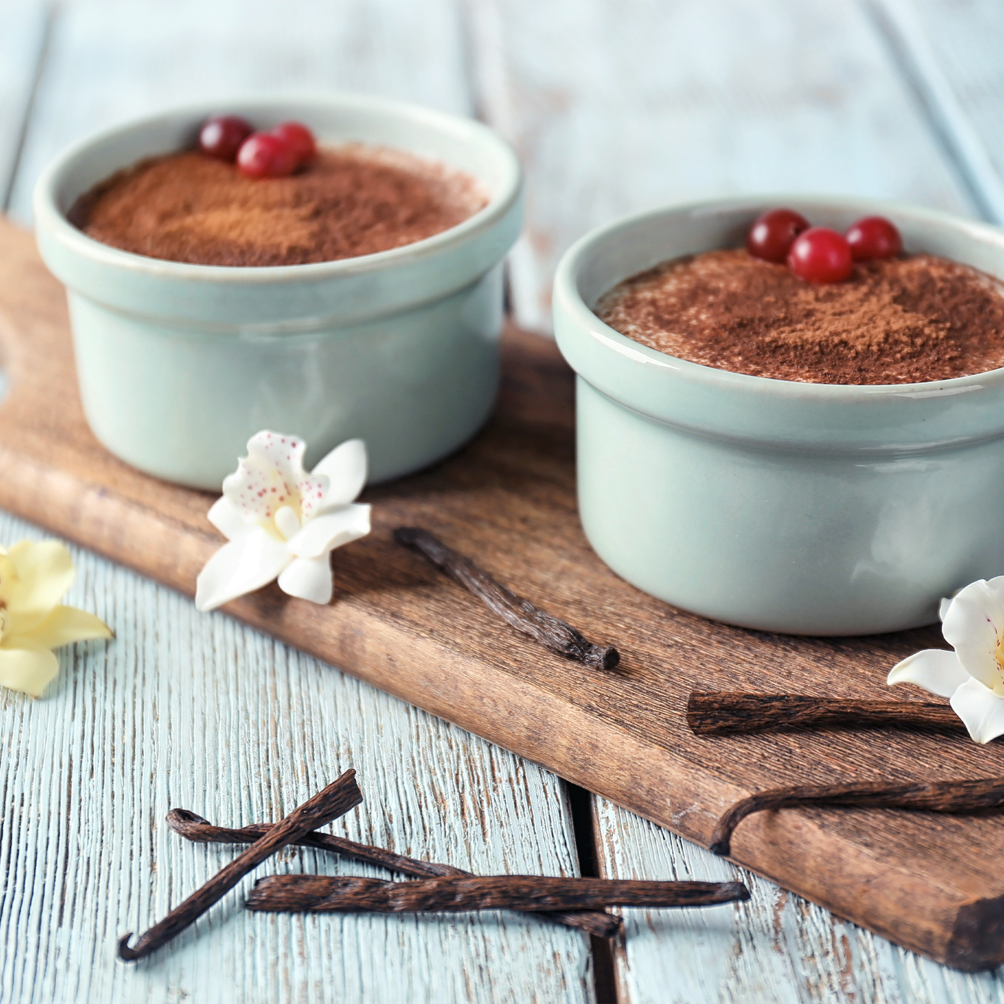 Two pale green ramekins filled with cocoa-dusted dessert and topped with red berries, arranged on a wooden board with vanilla beans and white flowers on a light blue wooden table.